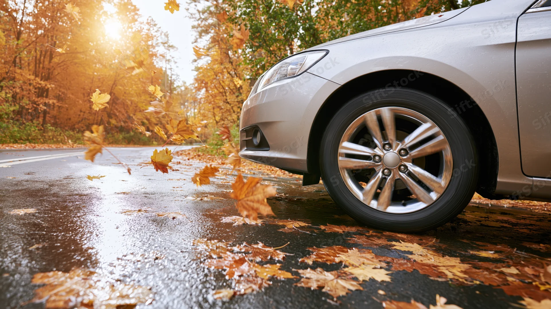 Silver sedan driving on wet autumn road with falling leaves swirling around it