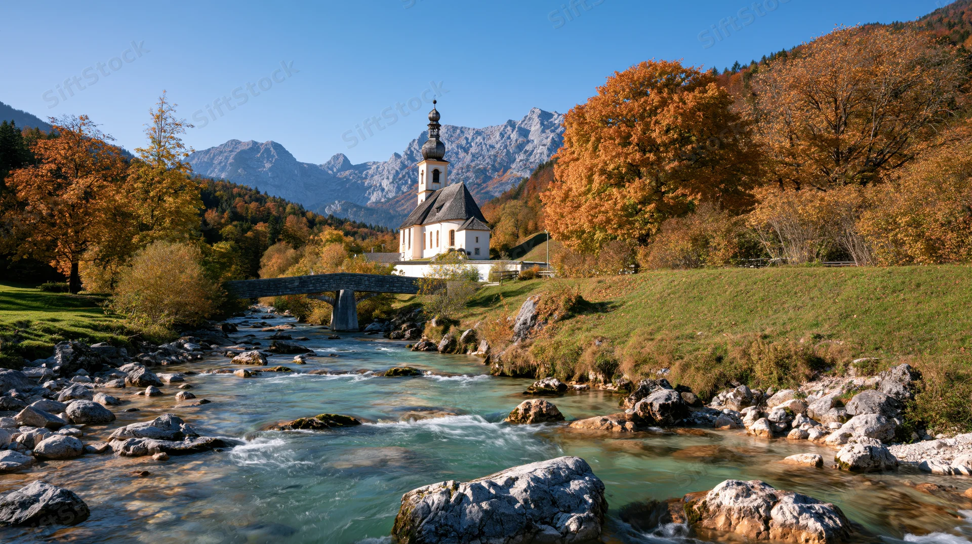 Small Bavarian village church with onion dome steeple nestled beside a flowing river in autumnal alpine landscape