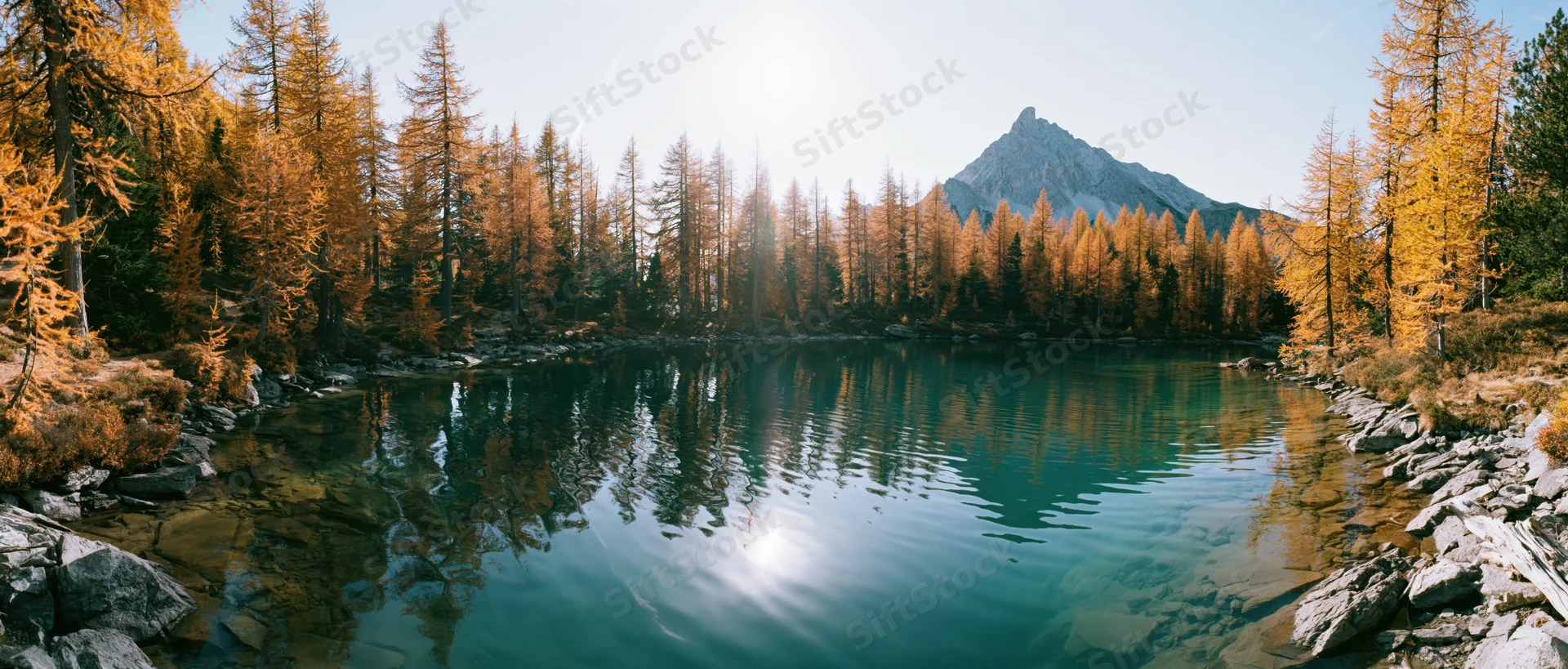 Alpine lake with clear turquoise water reflecting golden autumn trees and a mountain peak under sunlight