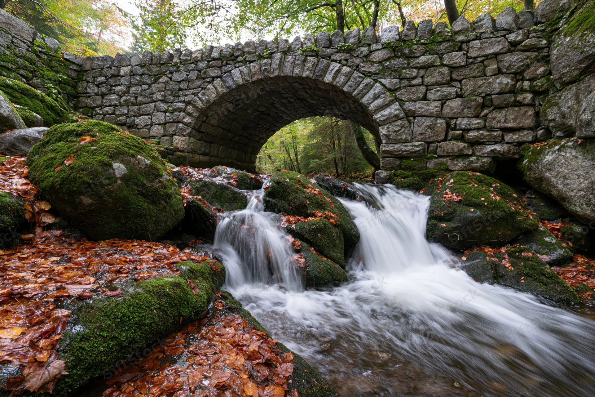 Small cascading waterfall flows over mossy rocks beneath a stone arch bridge in a forest during autumn
