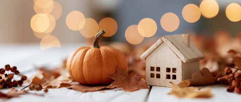 Small orange pumpkin and wooden house figurine on a white table surrounded by autumn leaves and warm bokeh lights