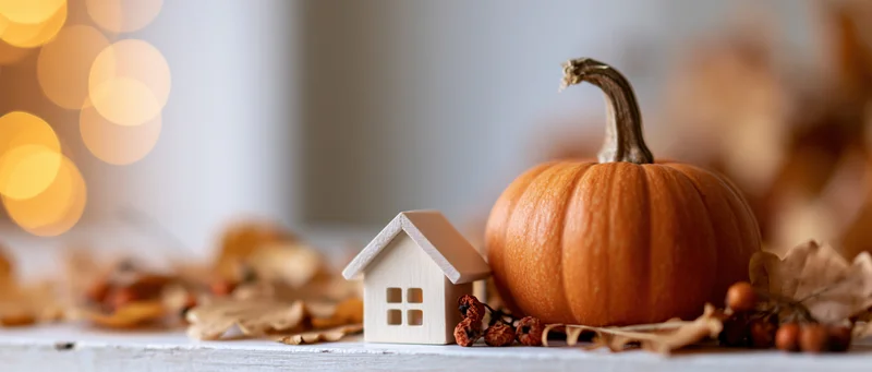 Small orange pumpkin and wooden house figurine arranged with autumn leaves and berries on a table with warm bokeh lights in the background