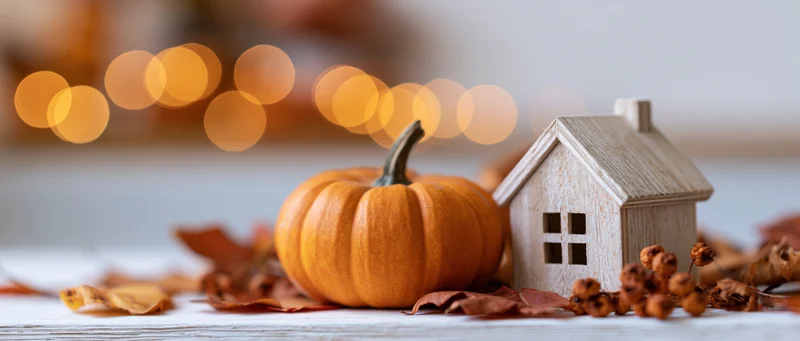 Small orange pumpkin and wooden house figurine on a tabletop with autumn leaves and bokeh lights in the background