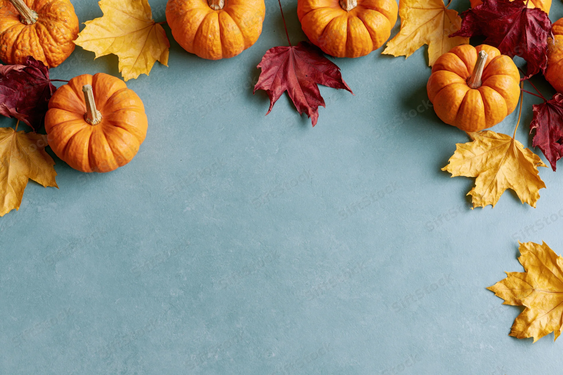 Small orange pumpkins and dried maple leaves arranged around the edges of a blue surface, leaving central copyspace