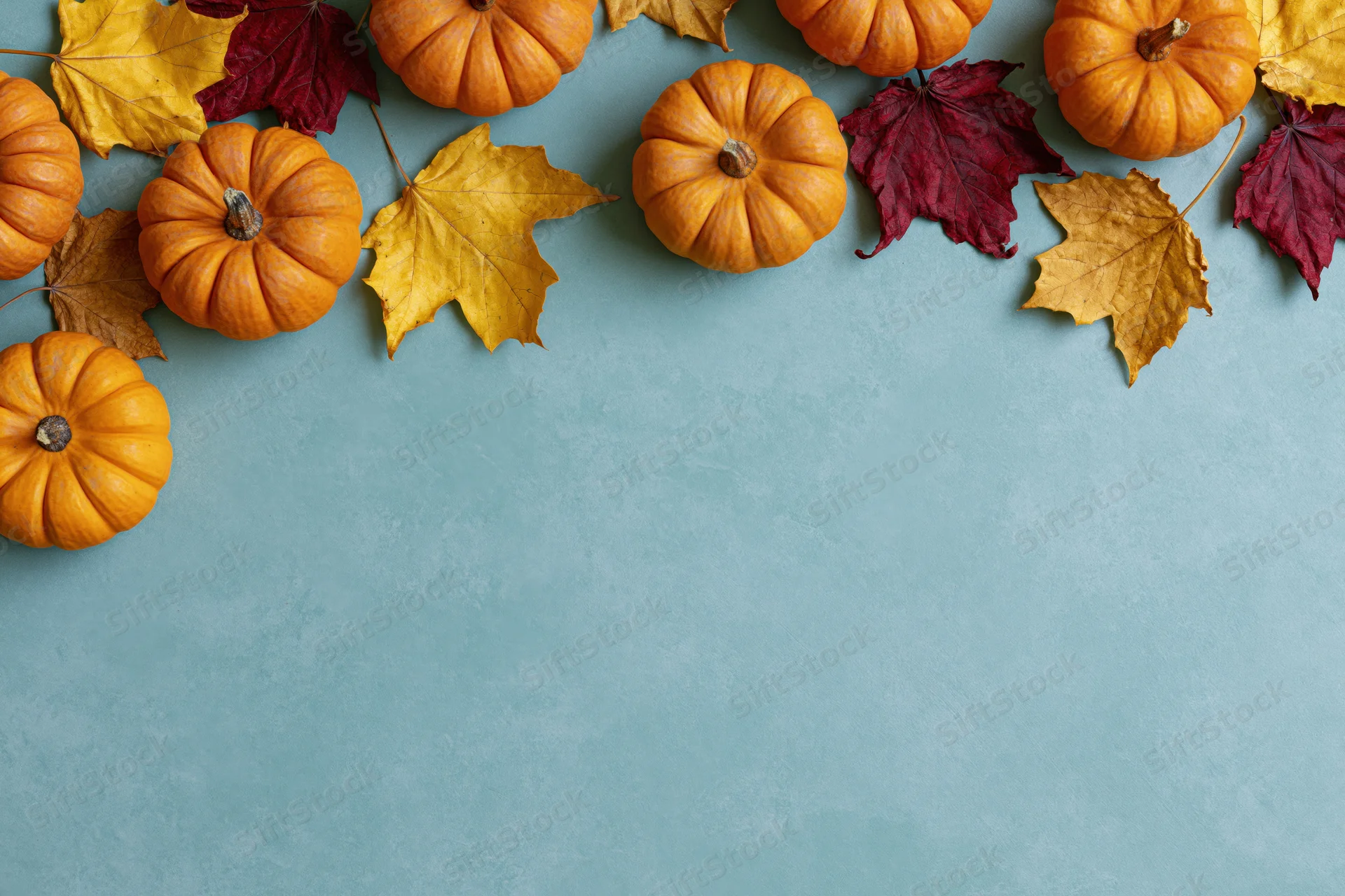 Small orange pumpkins and dried maple leaves arranged along the top of a teal surface, leaving copyspace below