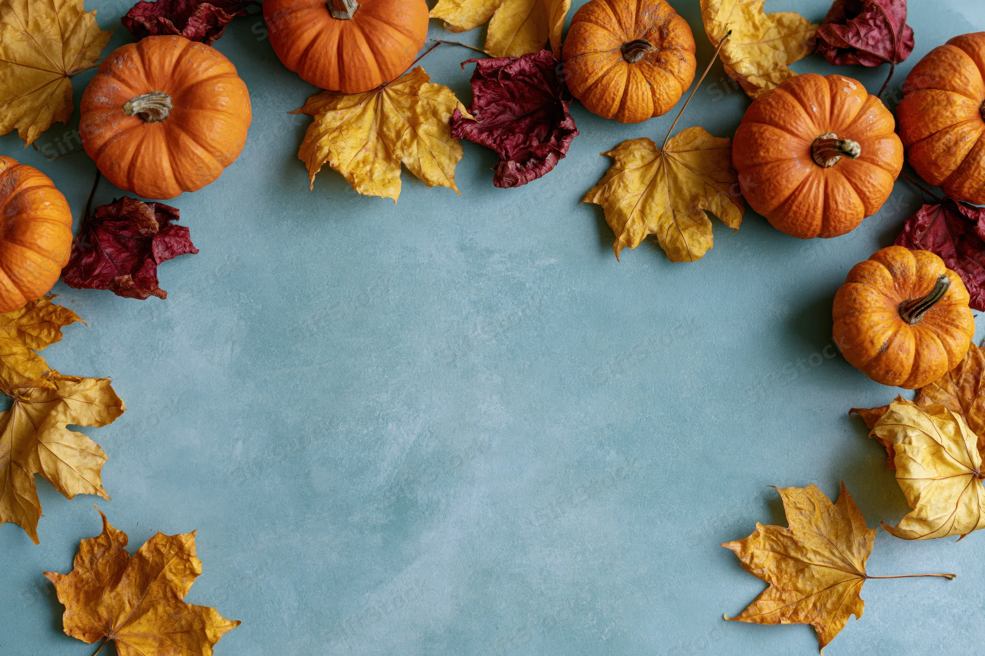 Small orange pumpkins and dried maple leaves arranged in a border around a blue surface with copyspace in the center