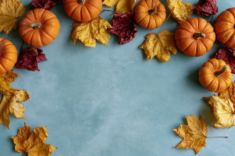 Small orange pumpkins and dried maple leaves arranged in a border around a blue surface with copyspace in the center