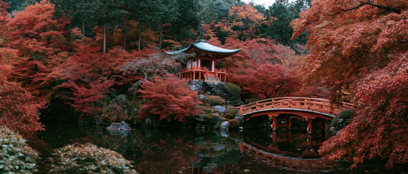 Small red japanese pagoda with green copper roof nestled among autumn maple trees beside a pond with a wooden bridge and reflections