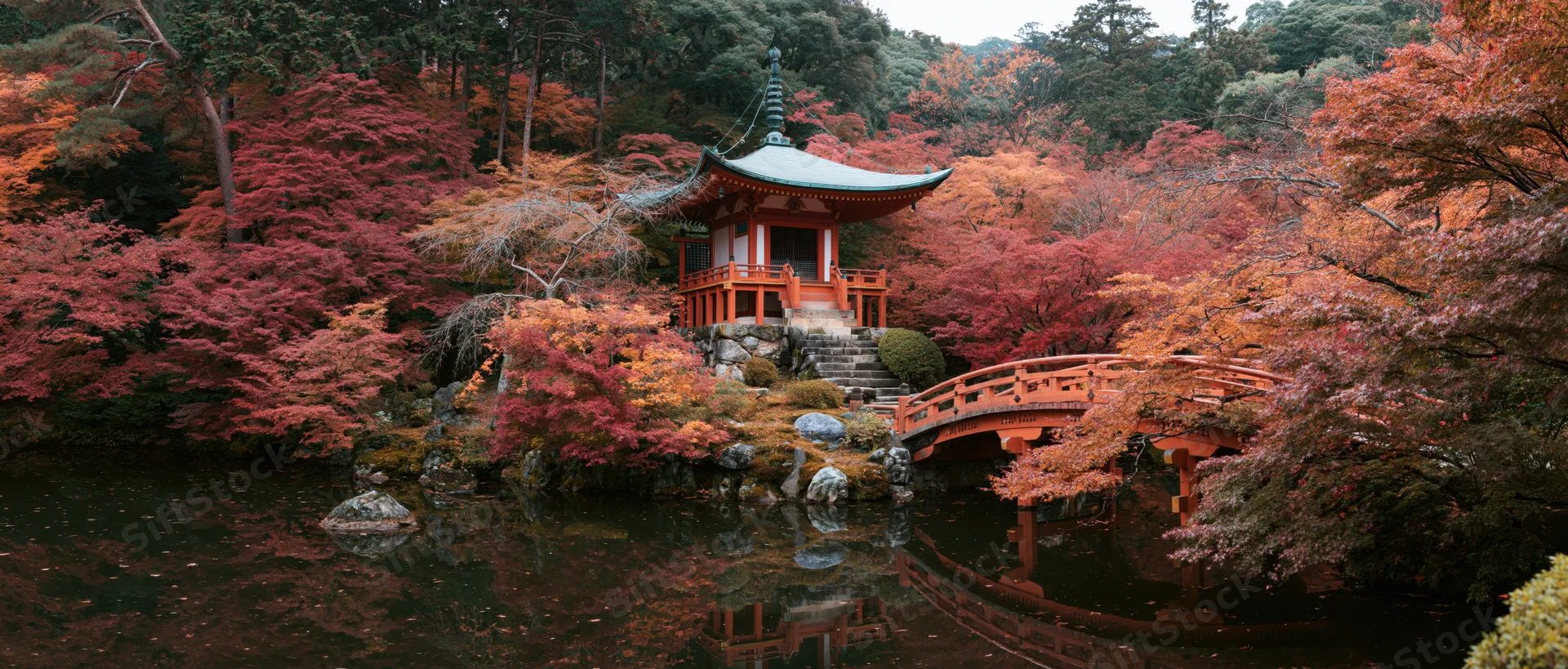 Small red japanese pagoda with green copper roof nestled among autumn maple trees beside a pond with a wooden bridge and reflections