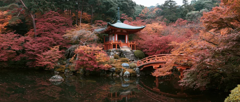Small red japanese pagoda with green copper roof nestled among autumn maple trees beside a pond with a wooden bridge and reflections