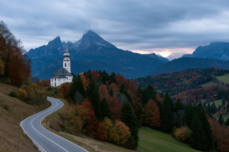 White baroque pilgrimage church with onion-dome tower on hillside beside winding road through autumn forest in alpine valley