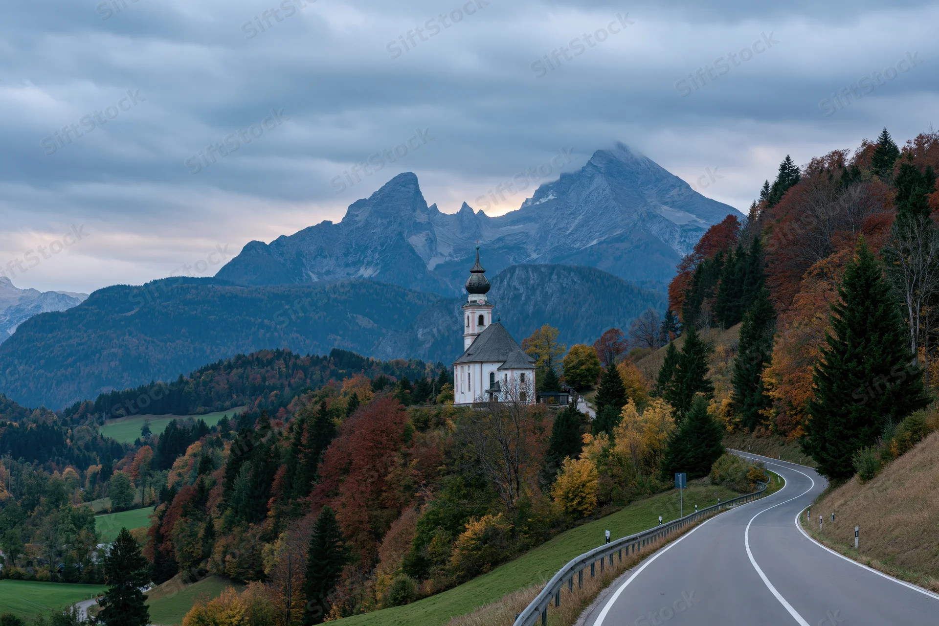 Winding road through autumn forest leading to white baroque pilgrimage church on hillside with mountains in background