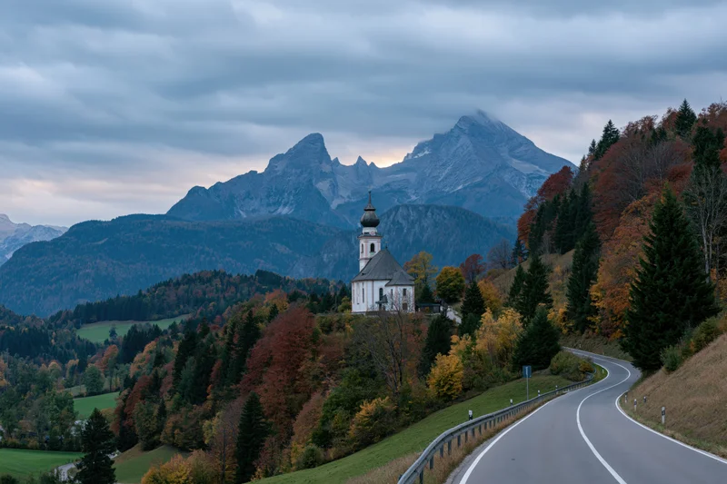 Winding road through autumn forest leading to white baroque pilgrimage church on hillside with mountains in background