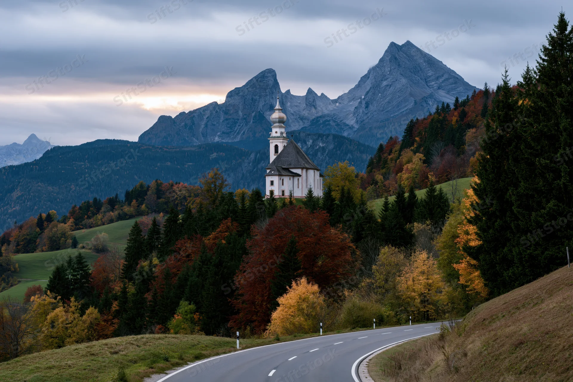 Small white baroque pilgrimage church with onion-dome tower perched on a hillside amid autumn forest and mountains, viewed from a winding road