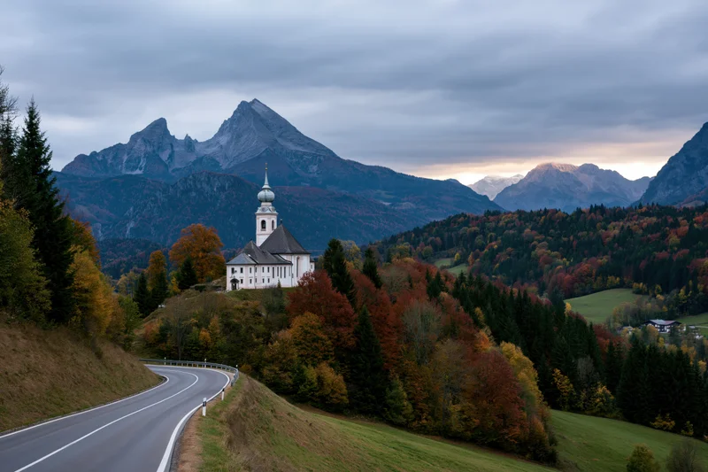 White baroque pilgrimage church with onion-dome tower on hillside overlooking winding road through autumn forested valley with mountains in background