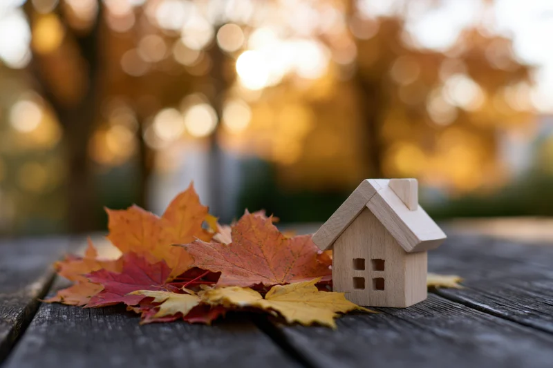 Small wooden house figurine resting on dark weathered wood table beside autumn maple leaves with blurred golden foliage background