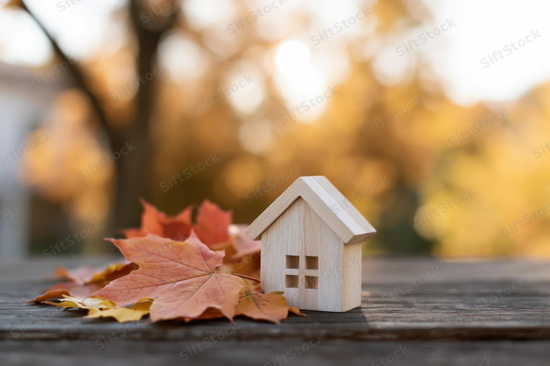 Small wooden house figurine on dark weathered table surrounded by autumn maple leaves with blurred golden foliage background
