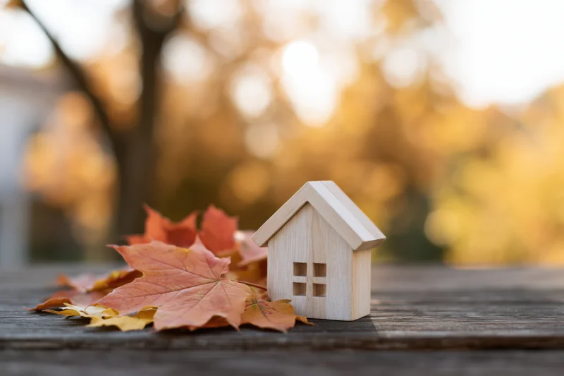 Small wooden house figurine on dark weathered table surrounded by autumn maple leaves with blurred golden foliage background