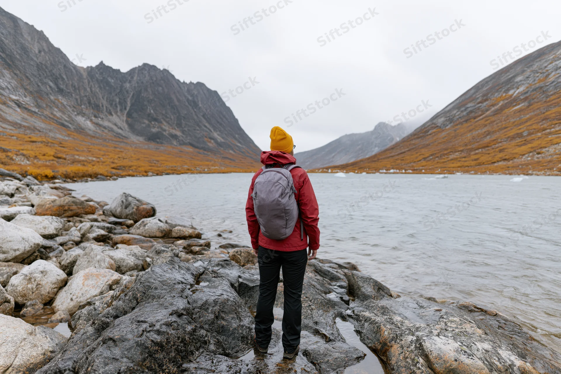 Woman hiker with backpack standing on rocky lakeshore facing mountains in autumn landscape