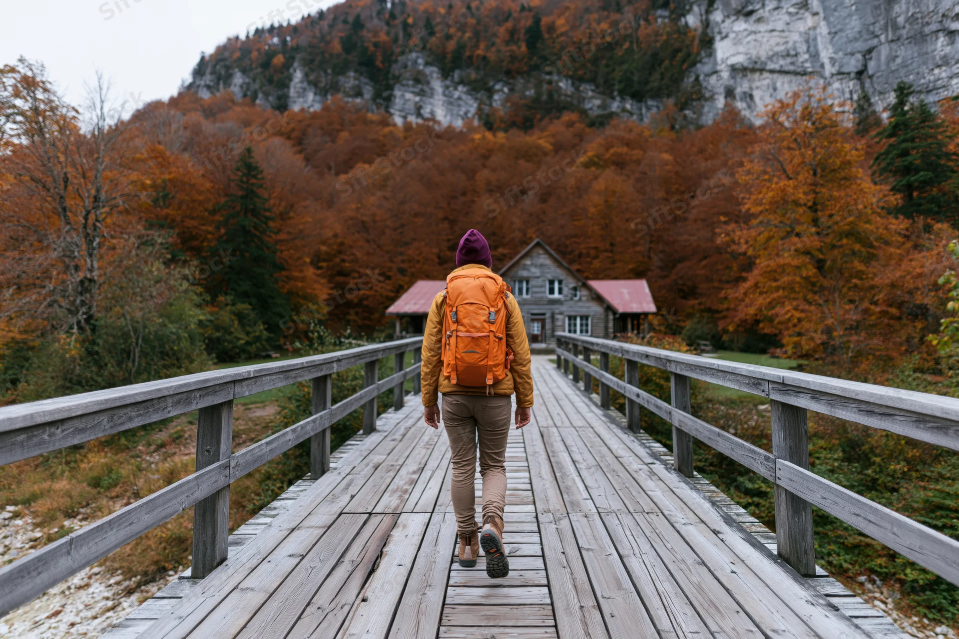 Hiker with orange backpack and purple hat walking away on wooden bridge toward cabin in autumn forest