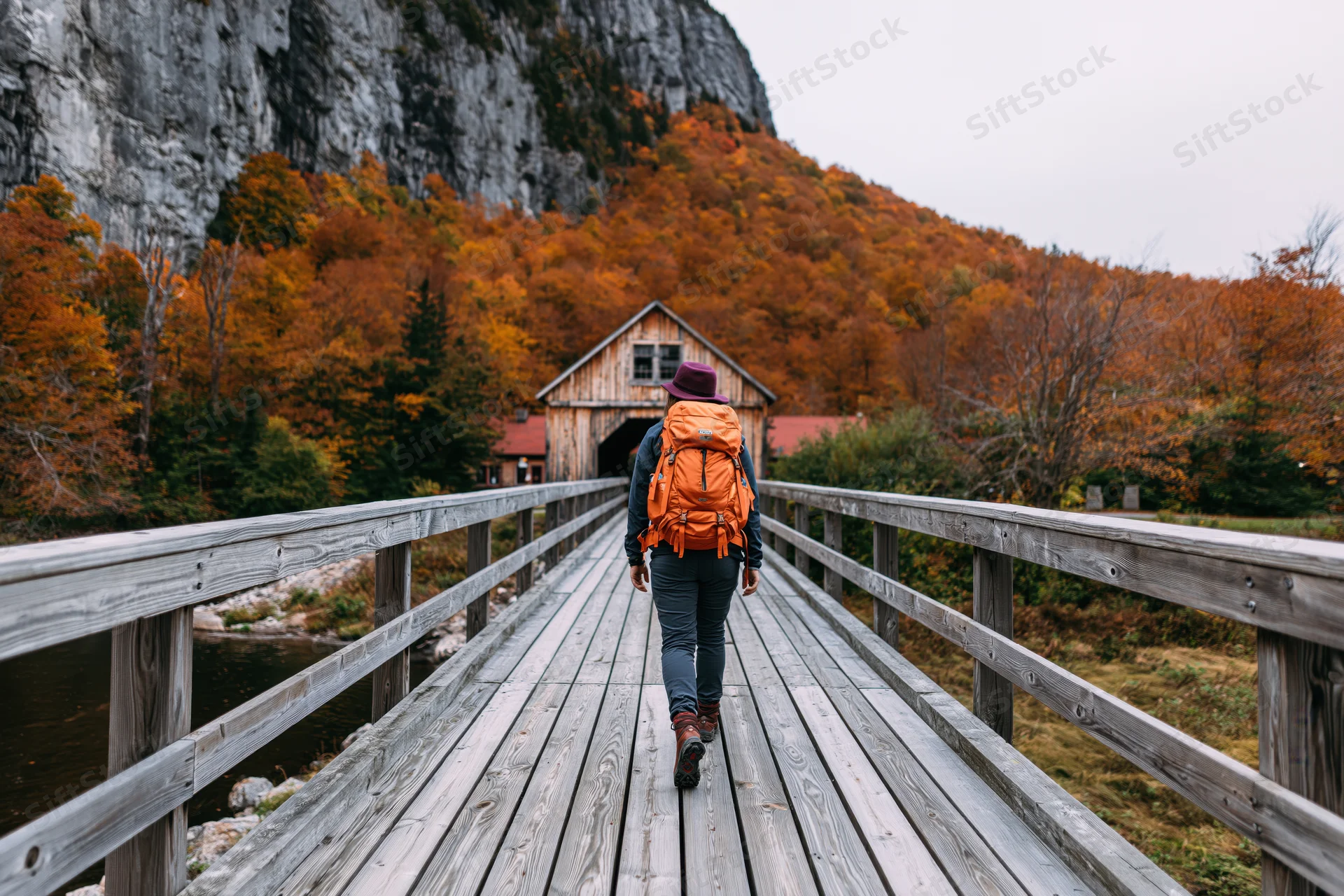 Woman with orange backpack and purple hat walking away on wooden bridge toward cabin in autumn forest