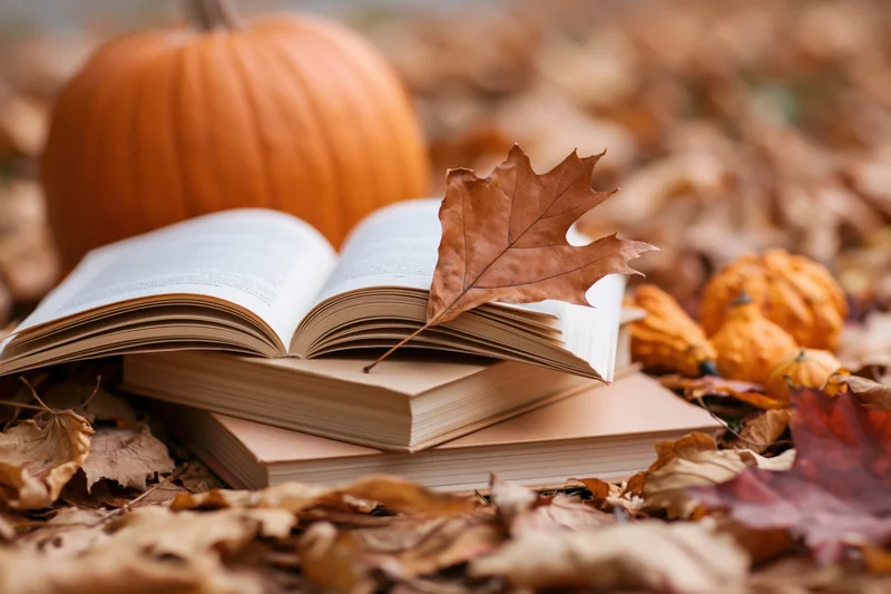 Stack of open and closed books resting among scattered autumn leaves with pumpkins on ground