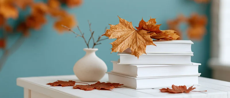 Stack of white hardcover books with dried maple leaves on a table beside a ceramic vase with a branch