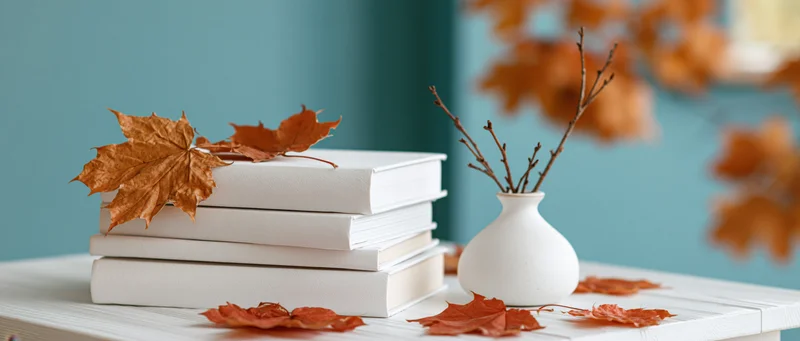Stack of white hardcover books with dried maple leaves on a wooden table beside a ceramic vase with branches