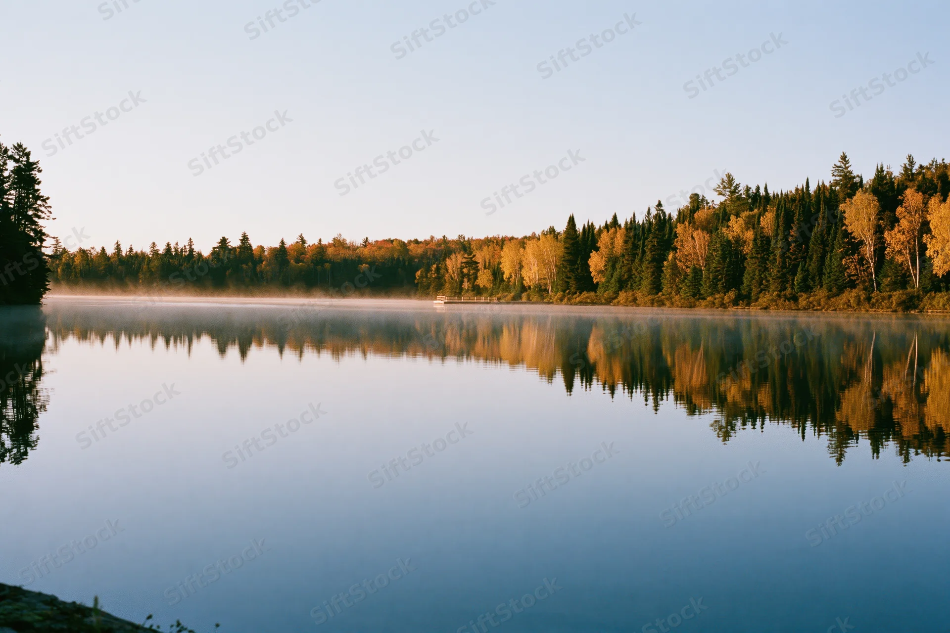 Calm lake reflecting autumn forest at early morning with mist drifting over water