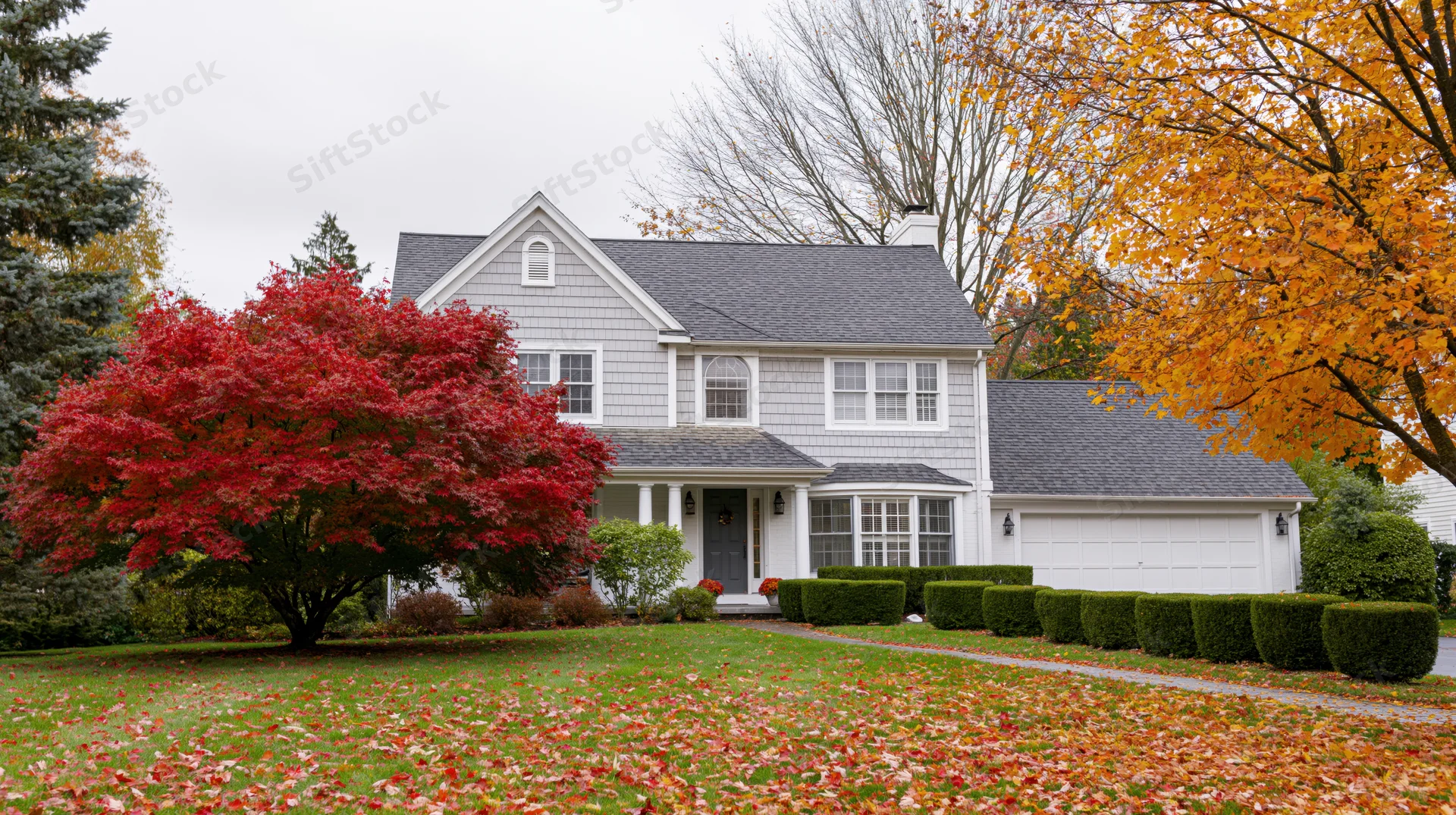Suburban two-story house with grey shingle roof and white siding surrounded by autumn trees and fallen leaves on the lawn