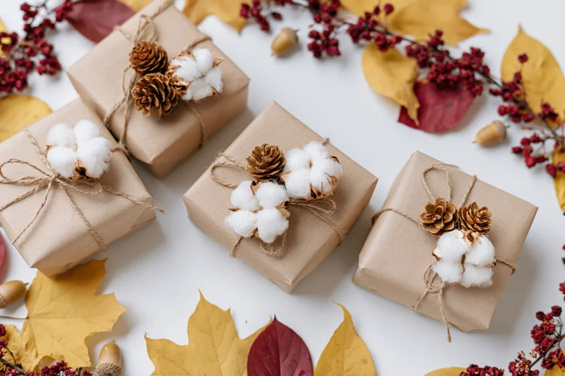Four kraft paper gift boxes wrapped with twine and decorated with pinecones and cotton, arranged on a white surface with autumn leaves and berries around