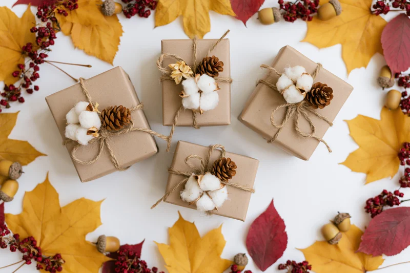 Four kraft paper gift boxes tied with twine and decorated with pinecones and cotton, arranged on a white surface surrounded by autumn leaves and berries