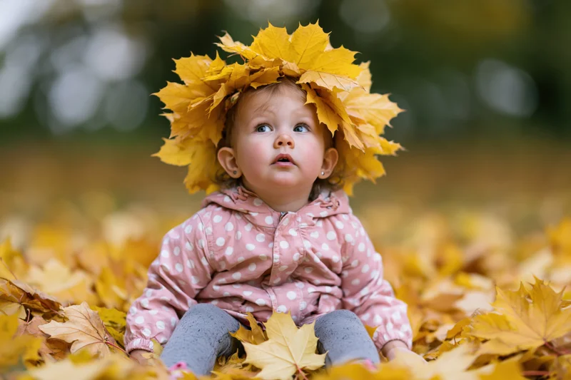 Toddler girl sitting in a pile of fallen autumn maple leaves wearing a leaf crown and pink polka dot jacket