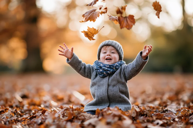 Toddler sitting on fallen autumn leaves in a park, arms raised, smiling, surrounded by falling foliage