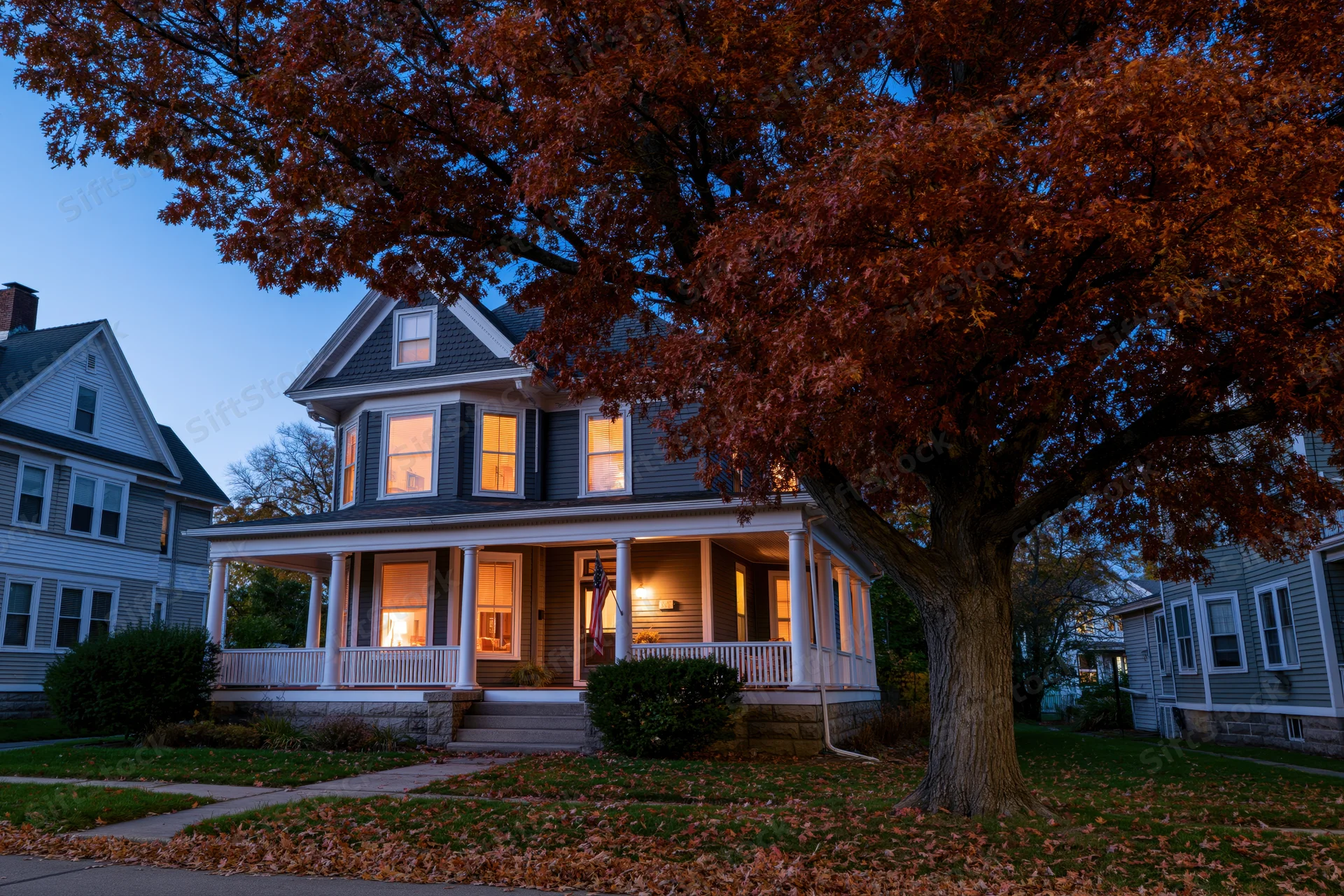 Victorian two-story house with wraparound porch and autumn tree in front yard at twilight