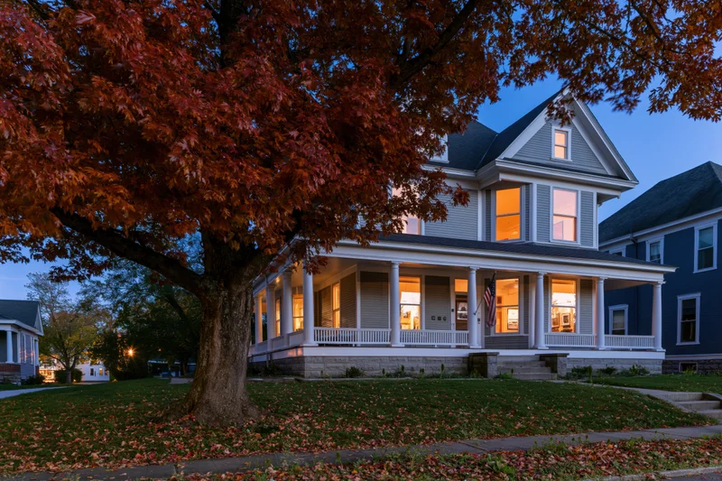 Victorian two-story house with wraparound porch and autumn tree in front yard at twilight