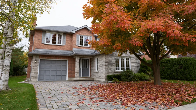 Two-story brick suburban house with grey roof and attached garage, surrounded by autumn foliage and fallen leaves on the driveway
