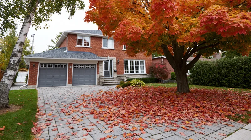 Two-story brick suburban house with grey roof and attached garage, surrounded by autumn foliage and fallen leaves on the driveway and lawn