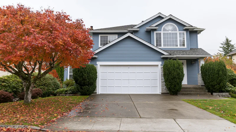 Two-story suburban house with blue-grey vinyl siding and white garage door, front entrance with steps and porch, surrounded by autumn trees and shrubs on a residential street