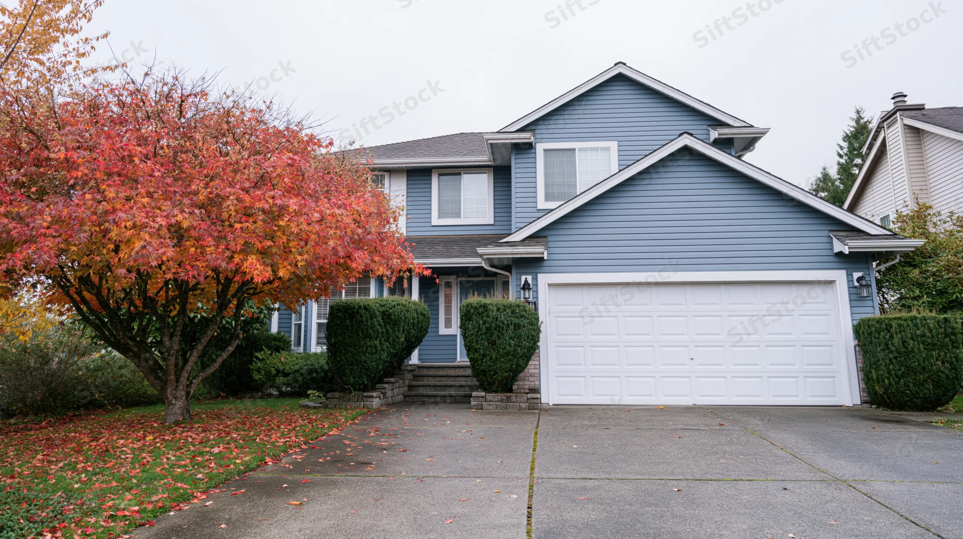 Blue-grey two-story suburban house with white garage and autumn tree in front yard