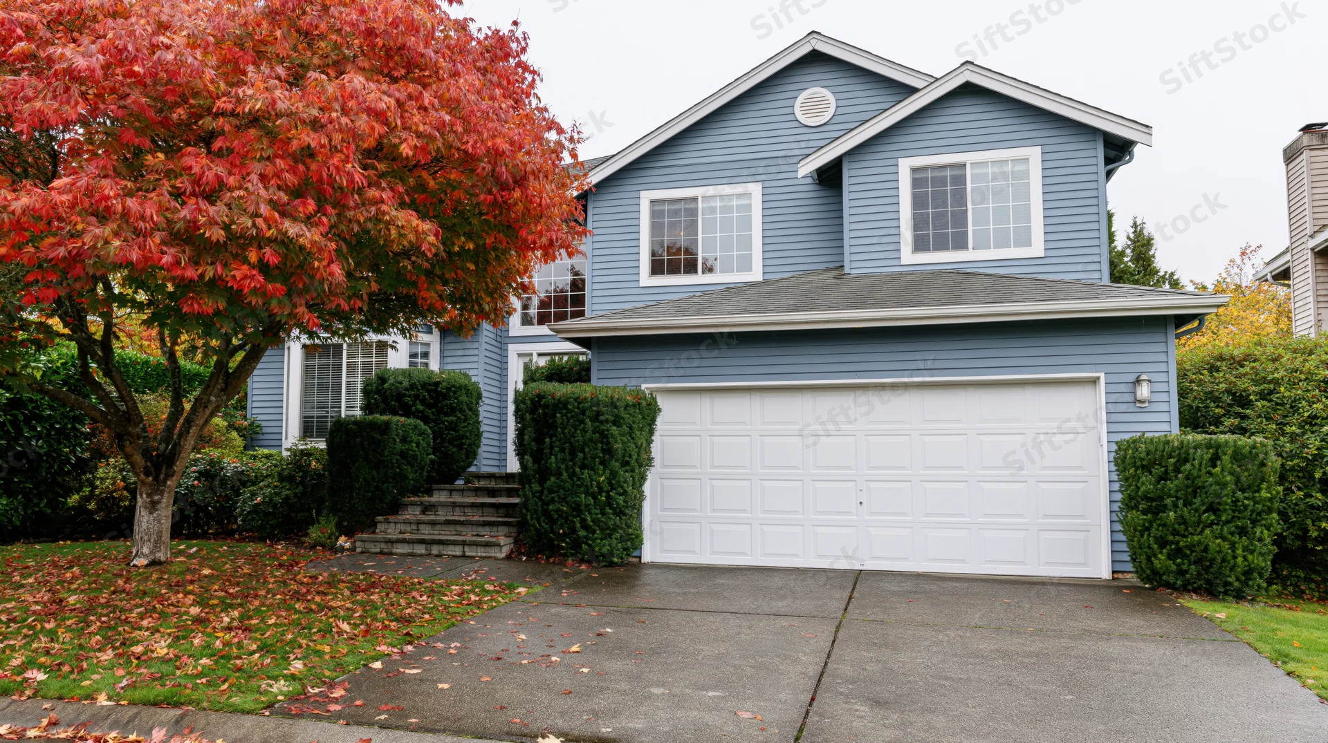 Blue-grey two-story suburban house with white garage and red-leafed tree in front yard during autumn