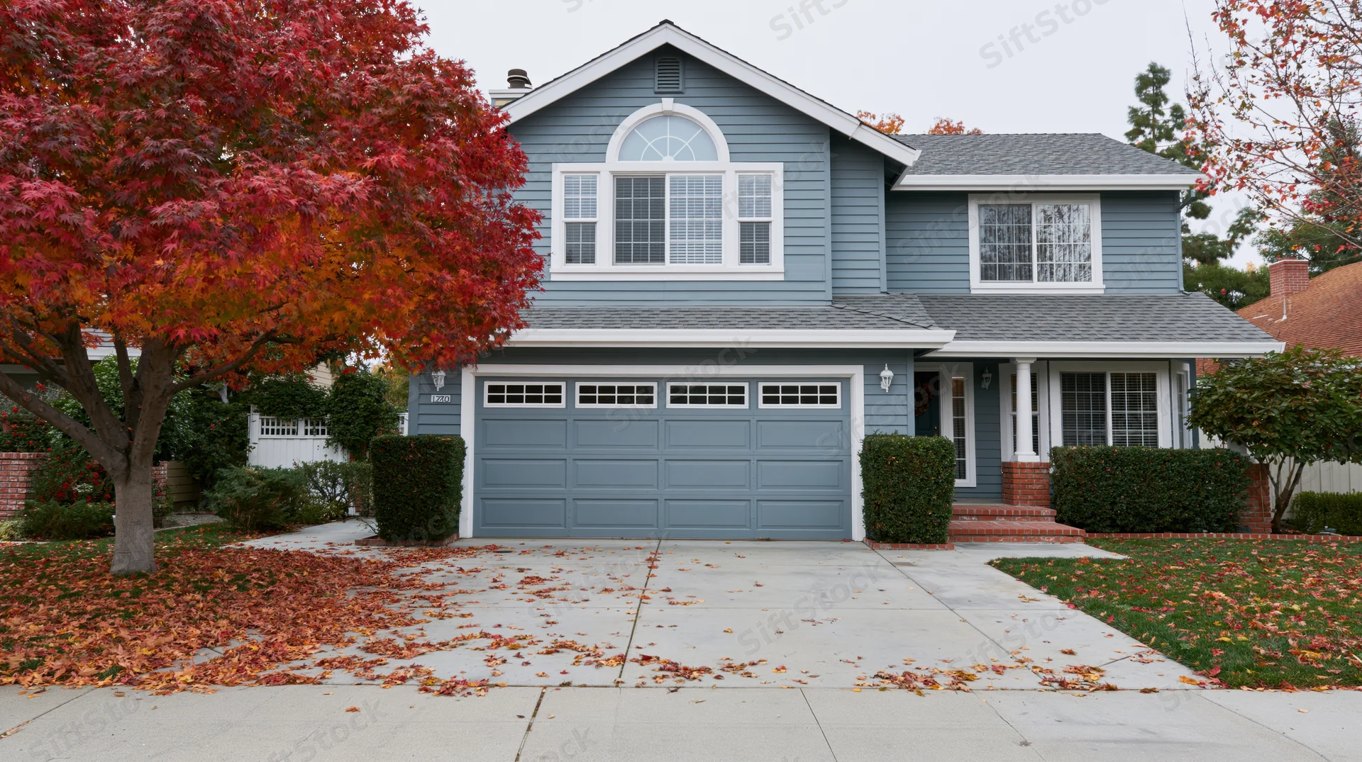 Two-story suburban house with grey-blue vinyl siding and white trim, front yard covered in autumn leaves, surrounded by trees and hedges
