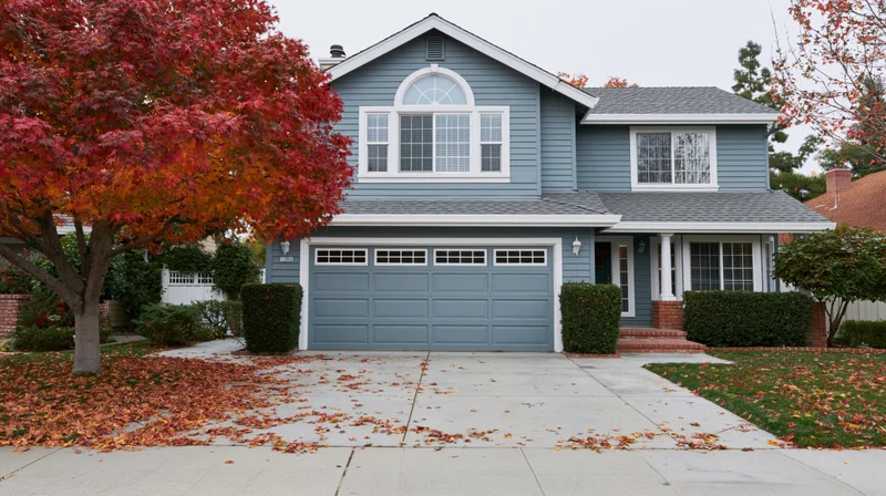 Two-story suburban house with grey-blue vinyl siding and white trim, front yard covered in autumn leaves, surrounded by trees and hedges