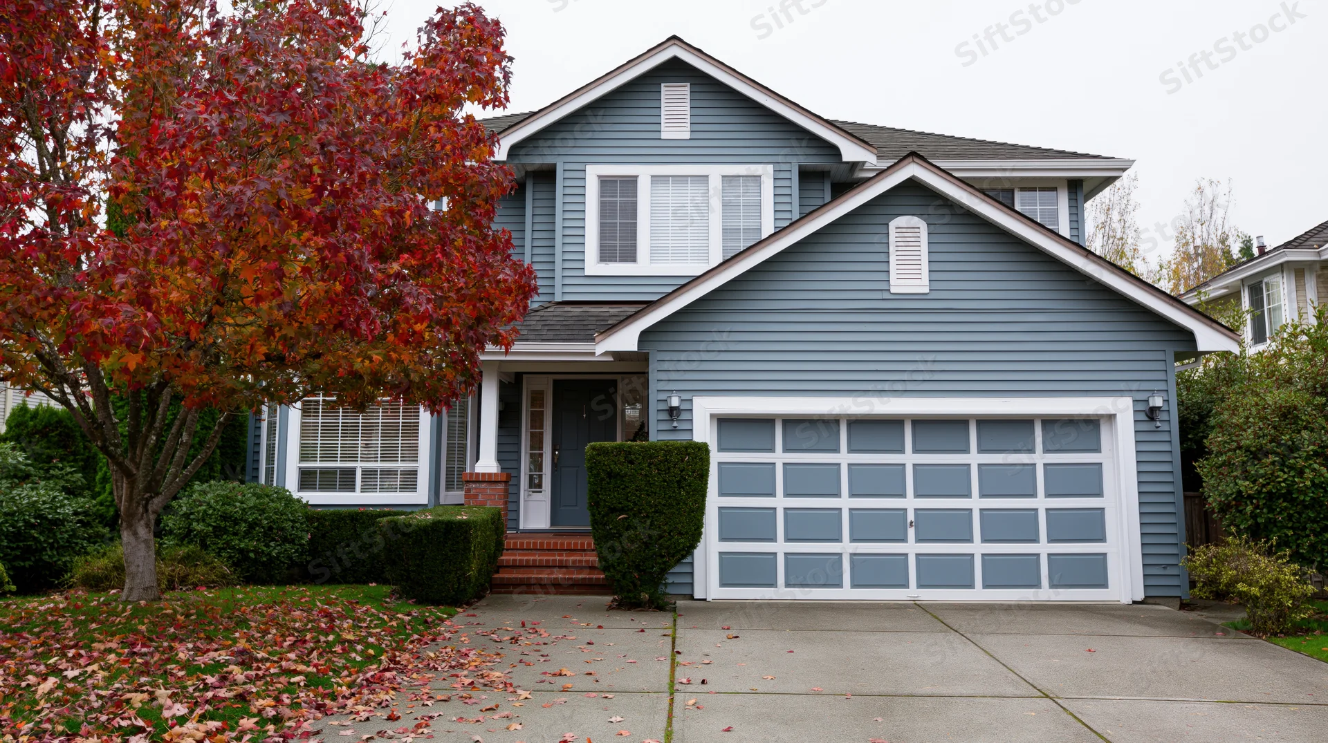 Two-story suburban house with grey-blue vinyl siding and white trim, front view showing garage, porch, and autumn tree with red leaves on lawn and driveway