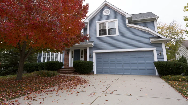 Two-story suburban house with grey-blue vinyl siding and white trim in autumn, surrounded by red-leafed tree and fallen leaves on driveway