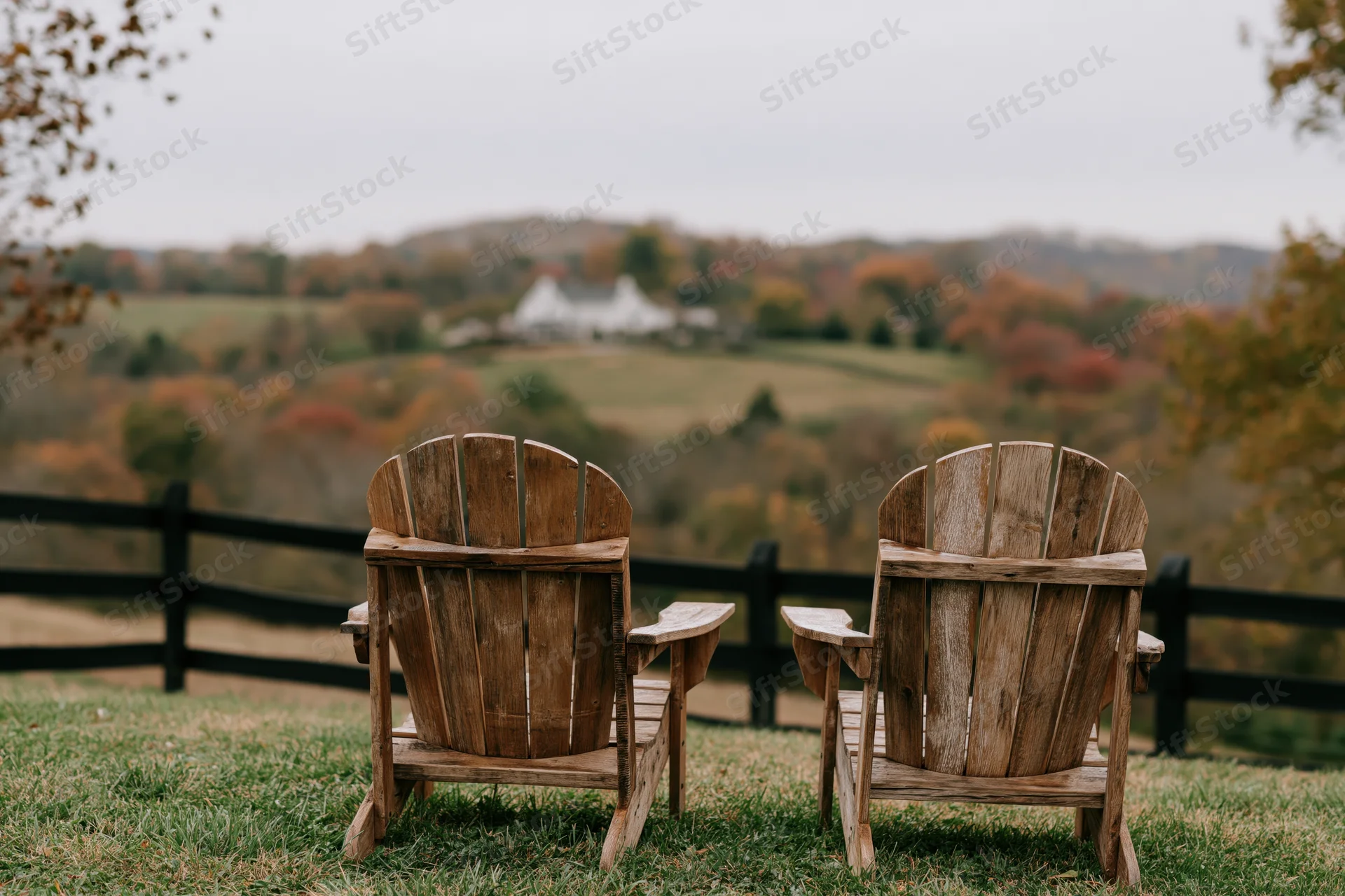 Two wooden adirondack chairs side by side on a grassy hill overlooking a rural autumn landscape with distant white farmhouse and colorful trees