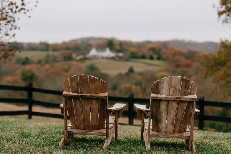 Two wooden adirondack chairs side by side on a grassy hill overlooking a rural autumn landscape with distant white farmhouse and colorful trees