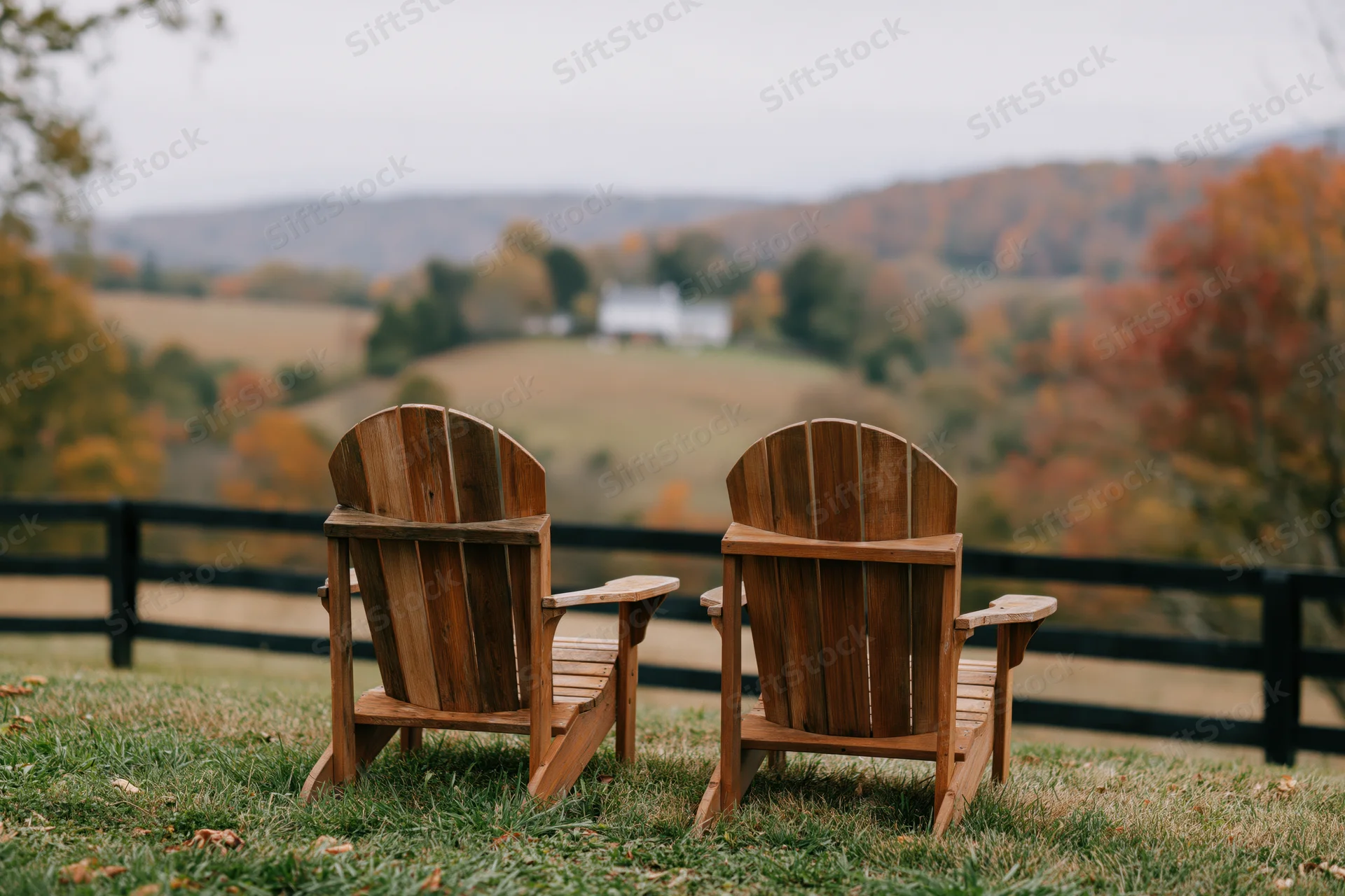 Two empty wooden adirondack chairs side by side on a grassy hill overlooking a rural autumn landscape