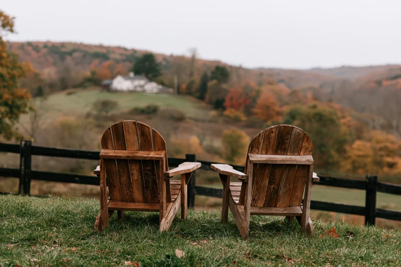 Two empty wooden adirondack chairs facing a scenic autumn landscape with rolling hills and a distant farmhouse