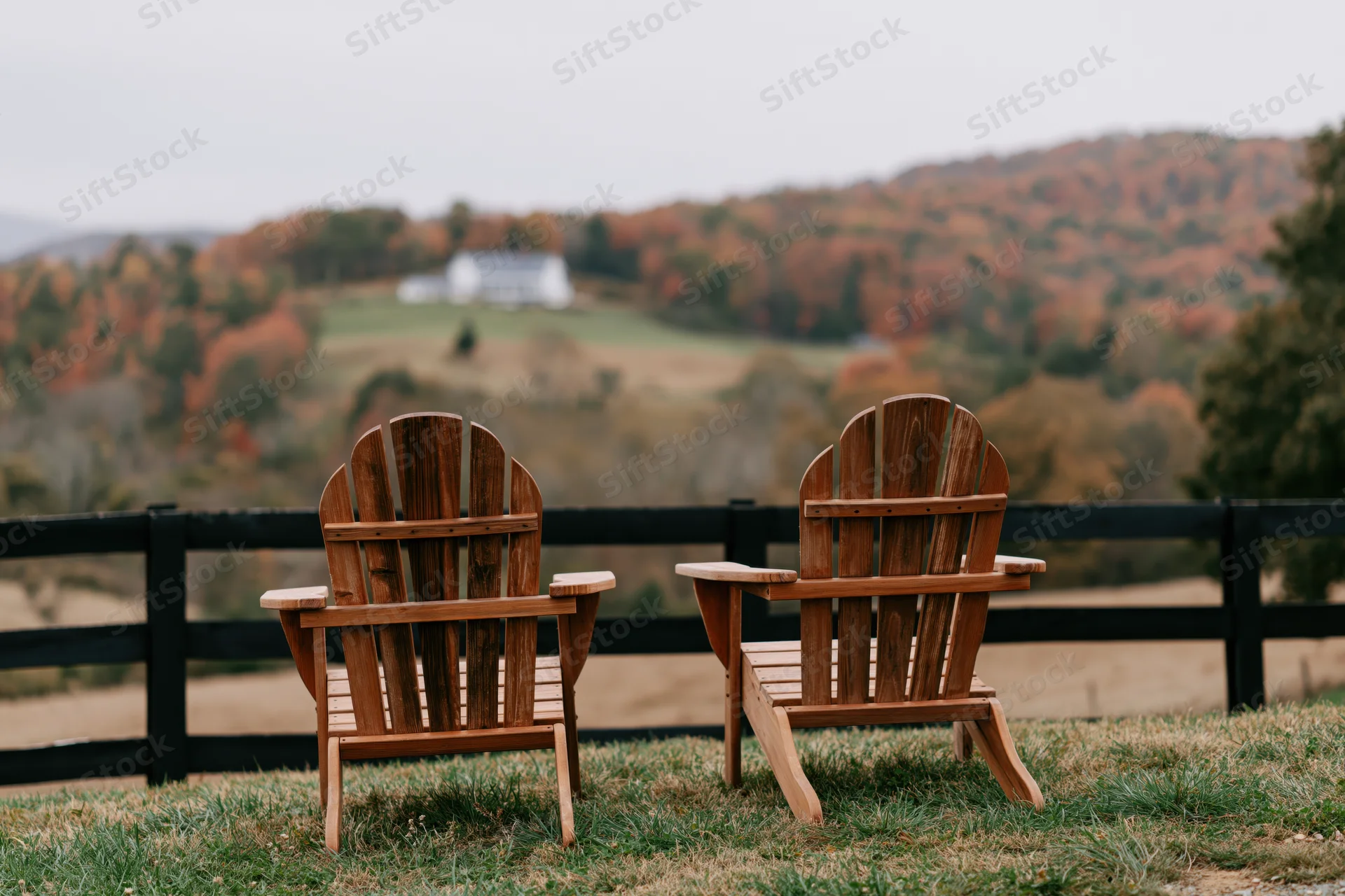 Two empty wooden adirondack chairs side by side on a grassy hill overlooking autumn hills and a distant white house